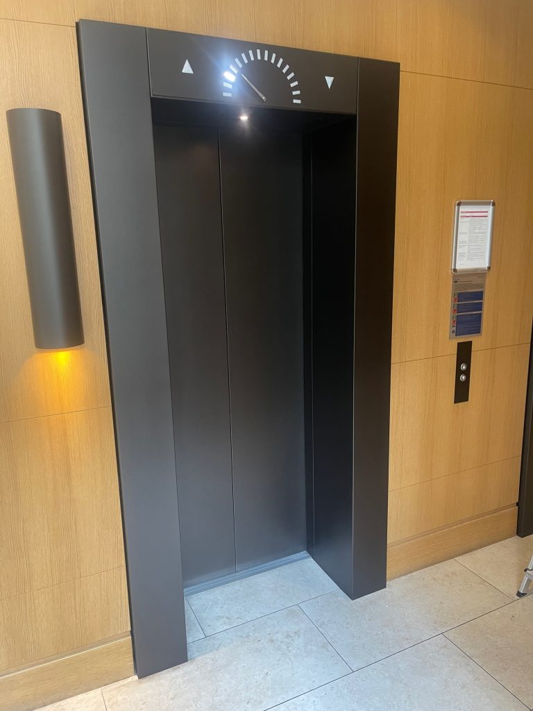 Dark sprayed lift door surround in a timber panelled lobby